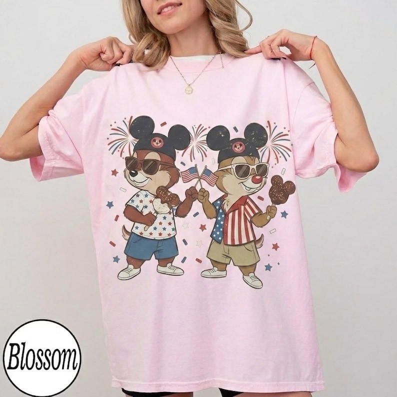 May include: A light pink t-shirt featuring a cartoon illustration of two chipmunks wearing sunglasses and Mickey Mouse hats. One chipmunk holds an American flag and the other holds a chocolate ice cream bar. Fireworks and stars are in the background.
