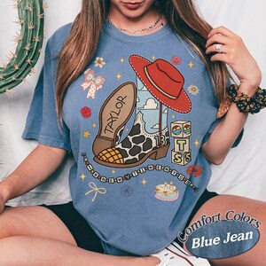 May include: A blue short-sleeve t-shirt featuring a graphic print of a cowboy boot with a cow print pattern, a red hat, a cake, and blocks with the letters T, S, and 13. The text Taylor and I KNEW YOU WERE TROUBLE are included.