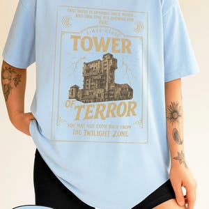 May include: Pale blue t-shirt featuring a gold graphic of the Hollywood Tower of Terror. The text on the shirt includes "TOWER OF TERROR" and "THE TWILIGHT ZONE". The shirt is made of a soft material.