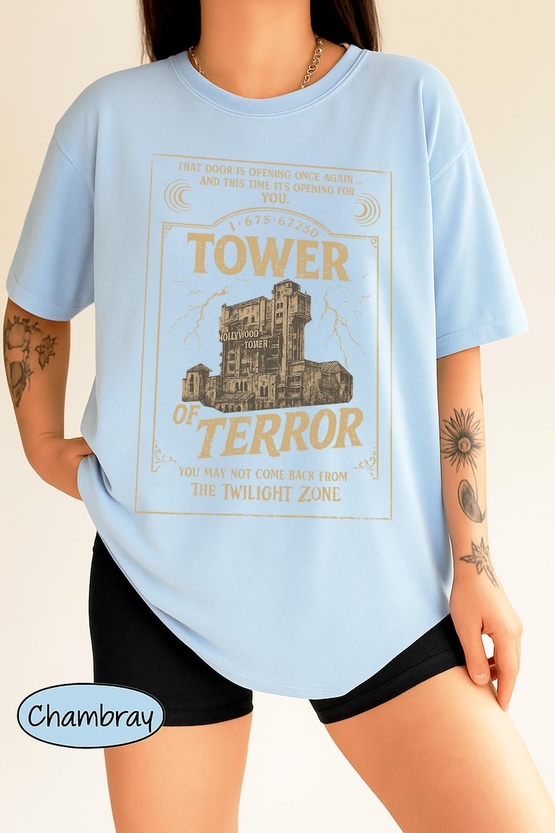 May include: Pale blue t-shirt featuring a gold graphic of the Hollywood Tower of Terror. The text on the shirt includes "TOWER OF TERROR" and "THE TWILIGHT ZONE". The shirt is made of a soft material.