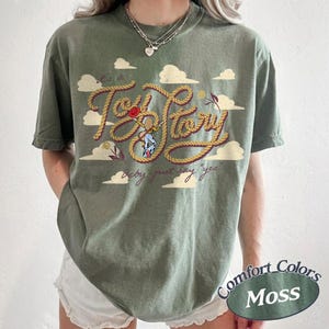 May include: An olive green t-shirt featuring a graphic print of a rope-style font that reads "it's a Toy Story" with a small illustration of a cowgirl character in the center and the text "baby just say yes" below, surrounded by white clouds.