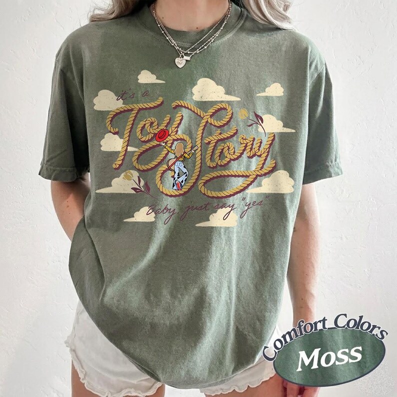 May include: An olive green t-shirt featuring a graphic print of a rope-style font that reads "it's a Toy Story" with a small illustration of a cowgirl character in the center and the text "baby just say yes" below, surrounded by white clouds.