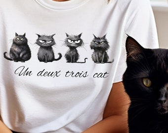 May include: White t-shirt with a graphic of four black cats with angry expressions and the text "Un deux trois cat". A black cat is next to the person wearing the shirt. The shirt is a casual, everyday wear item.