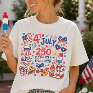 Puede incluir: Camiseta color crema con un gráfico colorido que celebra el 4 de julio y 250 años. El diseño incluye una bandera estadounidense, fuegos artificiales y otros símbolos patrióticos. La persona lleva unos pantalones cortos a cuadros verdes y blancos.