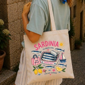 May include: Cream-colored tote bag with a colorful Sardinian design. The bag features illustrations of sardines, lemons, and a sailboat, with text that reads "Sardinia La Dolce Vita" and "Italian Summer". The bag has cream-colored straps.