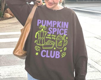 Può includere: Una felpa marrone con la scritta "PUMPKIN SPICE CLUB" in viola e verde. La grafica include una caffettiera, una torta e una tazza di pumpkin spice latte. La persona indossa jeans blu e una borsa marrone.