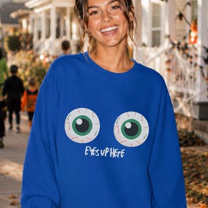 Op de afbeelding: Een koningsblauw sweatshirt met lange mouwen en een afbeelding van twee grote, doorbloede oogbollen met groene irissen in het midden op de borst. Onder de ogen staat de tekst "EYES UP HERE" gedrukt in een wit, handgeschreven lettertype.