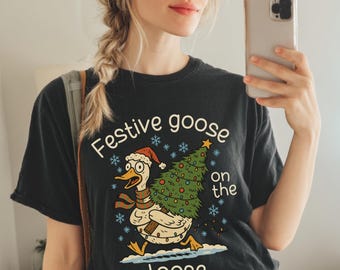 May include: Black t-shirt with the text "Festive goose on the loose" and a cartoon goose wearing a Santa hat and scarf, carrying a Christmas tree. The person in the image is wearing a red baseball cap and gray sweatpants.