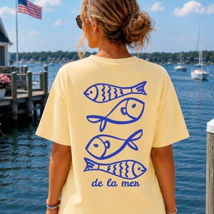 May include: A pale yellow short-sleeve t-shirt featuring a graphic print on the back with four stylized blue fish stacked vertically and the text de la mer written in blue script below the fish design, paired with blue denim shorts.
