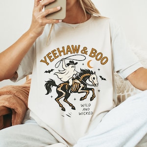 Könnte beinhalten: Cremefarbenes T-Shirt mit einem Halloween-Motiv. Das Design zeigt einen Geister-Cowboy, der ein Skelettpferd reitet, mit den Worten "Yeehaw & Boo" und "Wild and Wicked". Fledermäuse und ein Halbmond vervollständigen das Design.