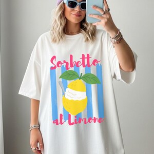 Puede incluir: Camiseta blanca con un gráfico de sorbete de limón. El diseño presenta un limón amarillo con sorbete, hojas verdes y el texto "Sorbetto al Limone" en rosa y azul. La camiseta tiene un corte holgado.