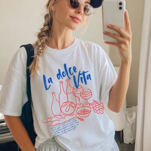 Puede incluir: Camiseta blanca con el texto "La dolce Vita" en azul y una ilustración roja de botellas de vino, copas y comida. La persona de la imagen lleva una gorra de béisbol azul marino y gafas de sol.
