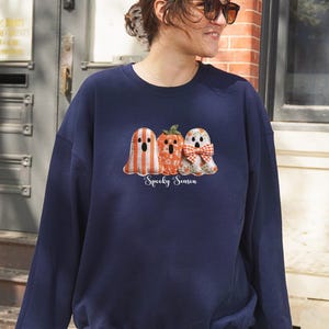 May include: Navy blue sweatshirt featuring a Halloween-themed graphic. The graphic includes three cartoon characters: a ghost with orange and white stripes, a pumpkin, and a ghost with a red and white bow. The text "Spooky Season" is below the characters.