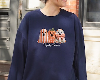 May include: A navy blue sweatshirt with a Halloween design. The graphic shows a ghost with orange and white stripes, a pumpkin, and a ghost with a red and white bow. The words "Spooky Season" are printed below the characters in a script font.