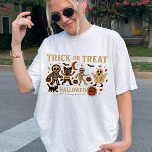 May include: White t-shirt with a Halloween-themed graphic. The design features the text "Trick or Treat" above illustrations of a skeleton, bear, mummy, and ghost, along with a black cat and pumpkins. The shirt has a crew neck and short sleeves.