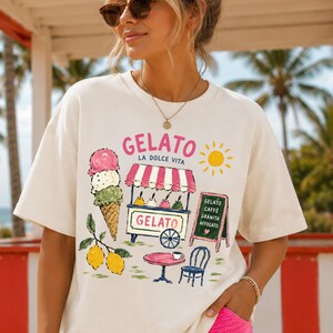 Puede incluir: Camiseta color crema con un gr&aacute;fico colorido de helado. El dise&ntilde;o incluye un cono de helado, un carrito de helado, limones y la palabra "Gelato" en rosa. Tambi&eacute;n hay un sol.