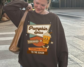Può includere: Una felpa marrone con la scritta "Pumpkin Spice" in un design retrò. Sotto l'insegna c'è un personaggio a forma di tazza di caffè dei cartoni animati. La felpa ha anche il testo "Coffee Club" e "'Tis the Season".