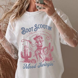 May include: White t-shirt with a graphic featuring a cowgirl, cacti, and the text "Boot Scootin' Mood Swingin'" in blue and pink. The shirt has a relaxed fit.