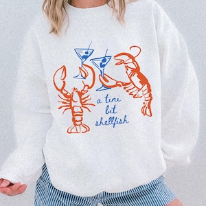 May include: White sweatshirt with a graphic design featuring two orange lobsters, two blue martini glasses, and the phrase "a tini bit shellfish" in blue script. The sweatshirt is paired with blue and white striped shorts.