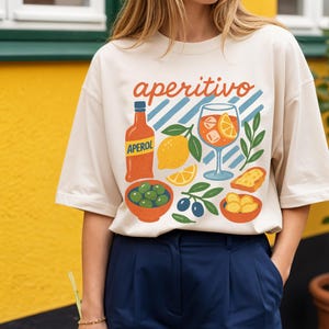 May include: Cream-colored t-shirt with the word "aperitivo" in red and orange script. The shirt features a colorful illustration of an Aperol bottle, a cocktail, olives, lemons, and bread. The model is wearing navy blue pants.
