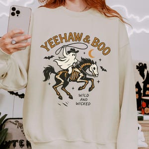 May include: Cream-colored sweatshirt with a Halloween-themed graphic. The design features a ghost riding a skeleton horse, holding a lasso, with the words "Yeehaw & Boo" and "Wild and Wicked". Bats and a crescent moon are also included.