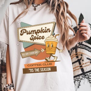May include: Off-white t-shirt with a vintage graphic design. The image shows a sign that says "Pumpkin Spice", a cartoon coffee cup, and the phrases "All Day, Every Day" and "'Tis The Season".