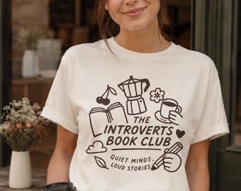 May include: A cream-colored short-sleeve t-shirt featuring a dark brown graphic print. The design includes illustrations of a book, coffee pot, cup, pencil, and cherry with the text: THE INTROVERTS BOOK CLUB, QUIET MINDS, LOUD STORIES.