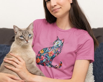 May include: A woman wearing a pink t-shirt with a floral cat design holds a light brown cat in her arms. The cat is looking to the right of the frame.
