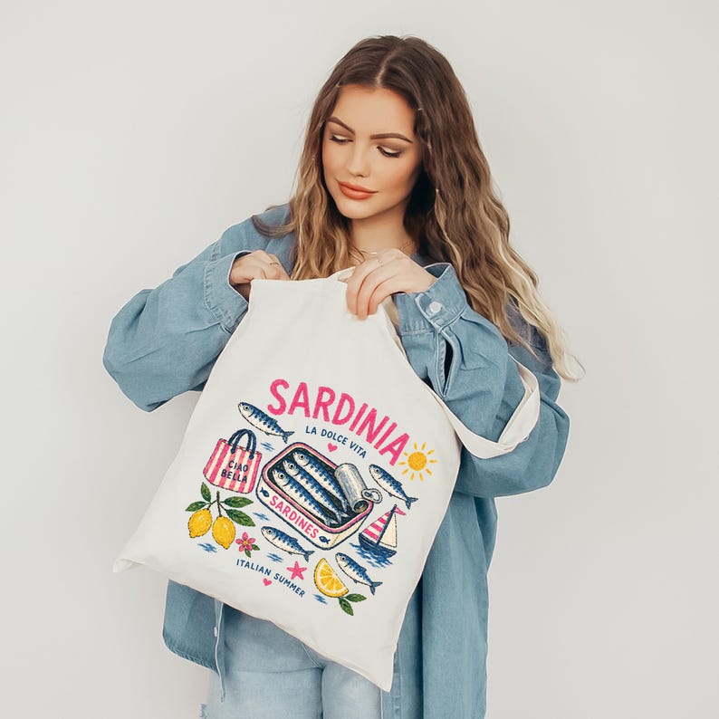 May include: Cream-colored tote bag with a colorful Sardinian design. The design includes the word "Sardinia" in pink, a can of sardines, fish, lemons, and a sailboat. The text "Italian Summer" is also included.