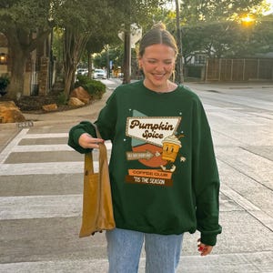 May include: A dark green sweatshirt with the words "Pumpkin Spice" and a cartoon coffee cup graphic. The sweatshirt also has the phrases "All Day, Every Day" and "Coffee Club 'Tis The Season". The person is holding a brown tote bag.