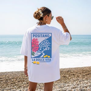 May include: White t-shirt featuring a blue rectangle with the word "POSITANO" above an illustration of a coastal town, pink flowers, and yellow lemons. The text "LA DOLCE VITA" is below the illustration. The shirt is worn on a beach.