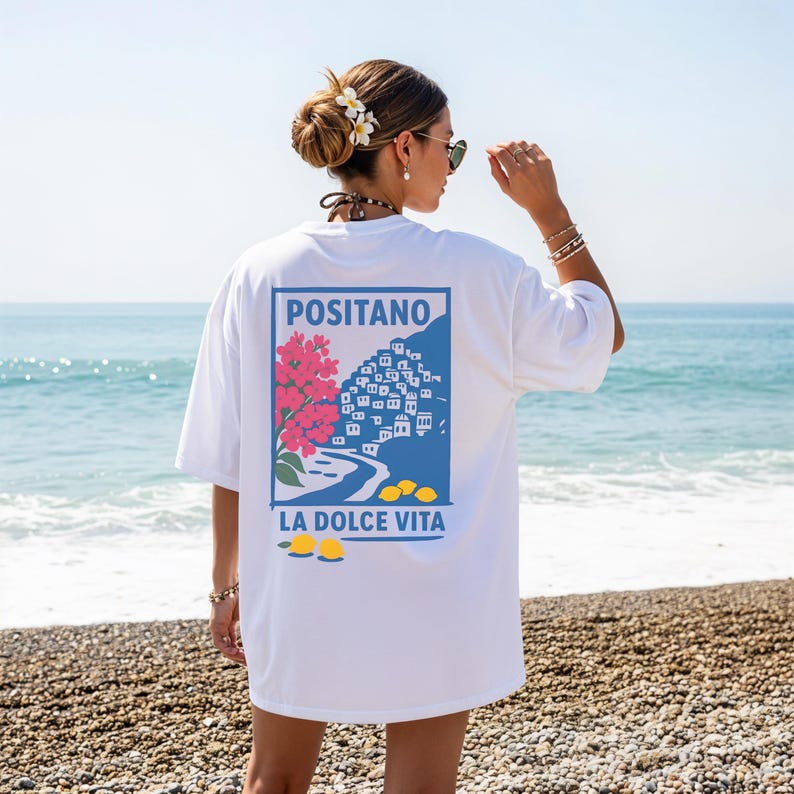 May include: White t-shirt featuring a blue rectangle with the word "POSITANO" above an illustration of a coastal town, pink flowers, and yellow lemons. The text "LA DOLCE VITA" is below the illustration. The shirt is worn on a beach.