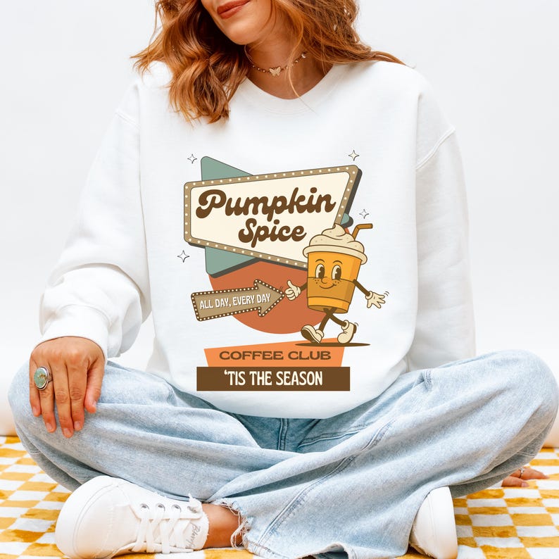 May include: White sweatshirt with a retro-style graphic. The graphic features a coffee cup character and the words "Pumpkin Spice Coffee Club" and "'Tis the Season". The design is in shades of brown, beige, and teal.