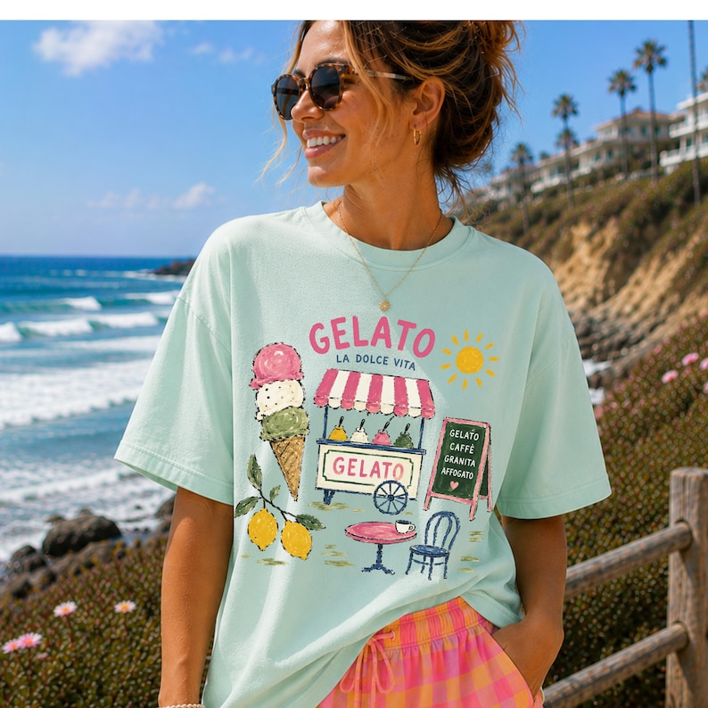 Puede incluir: Camiseta azul claro con un dise&ntilde;o de helado. El gr&aacute;fico incluye un cono de helado, un carrito de helado, limones y las palabras "Gelato La Dolce Vita". Una mujer con gafas de sol sonr&iacute;e en la imagen.