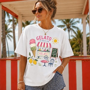 Puede incluir: Camiseta blanca con un dise&ntilde;o de helado colorido. El gr&aacute;fico incluye un cono de helado, un carrito de helados, limones y las palabras "Gelato La Dolce Vita". Un peque&ntilde;o letrero enumera los sabores de helado. La persona lleva gafas de sol.