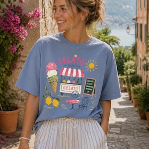 Puede incluir: Camiseta azul claro con un gr&aacute;fico de helado. El dise&ntilde;o incluye un cono de helado, un carrito de helado, limones y las palabras "Gelato La Dolce Vita". Una peque&ntilde;a pizarra enumera los sabores de helado. La camiseta se usa al aire libre.