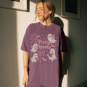 May include: A purple t-shirt featuring the phrase "Pretty Spooky" with illustrations of ghosts, hearts, bats, and bows. The design has a vintage aesthetic, ideal for Halloween.