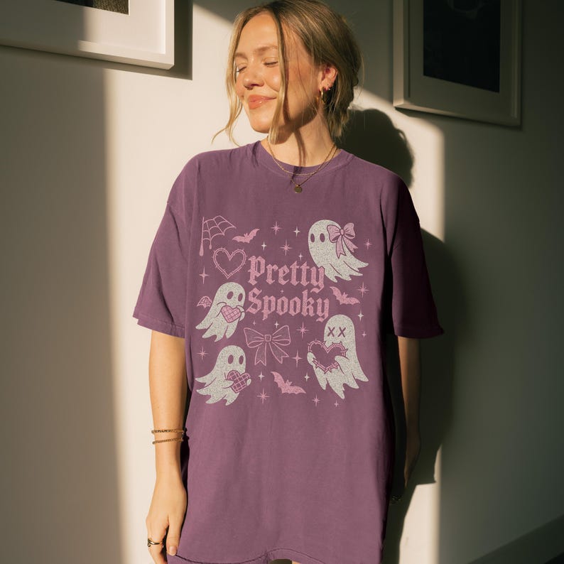 May include: A purple t-shirt featuring the phrase "Pretty Spooky" with illustrations of ghosts, hearts, bats, and bows. The design has a vintage aesthetic, ideal for Halloween.