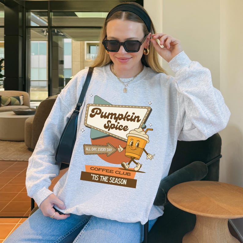 May include: A light gray sweatshirt with a retro-style graphic that reads "Pumpkin Spice" above a cartoon coffee cup character. The sweatshirt also has the phrases "All Day, Every Day" and "Coffee Club 'Tis The Season".