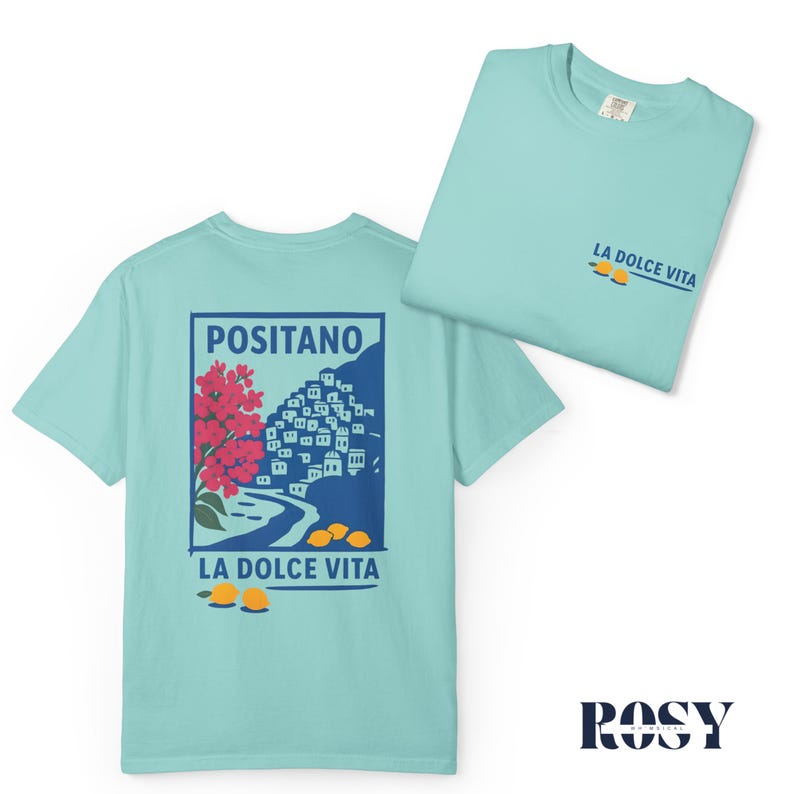 May include: Two light blue t-shirts. One shirt displays a graphic of Positano, Italy, with red flowers, white buildings, and yellow lemons. The text "POSITANO" and "LA DOLCE VITA" are included. The other shirt has "LA DOLCE VITA" and lemons.
