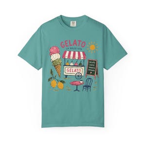 Puede incluir: Camiseta color turquesa con un dise&ntilde;o de helado. El gr&aacute;fico incluye un cono de helado, un carrito de helado con la palabra "Gelato", una mesa, una silla y el texto "La Dolce Vita". Tambi&eacute;n se incluye un sol y limones.