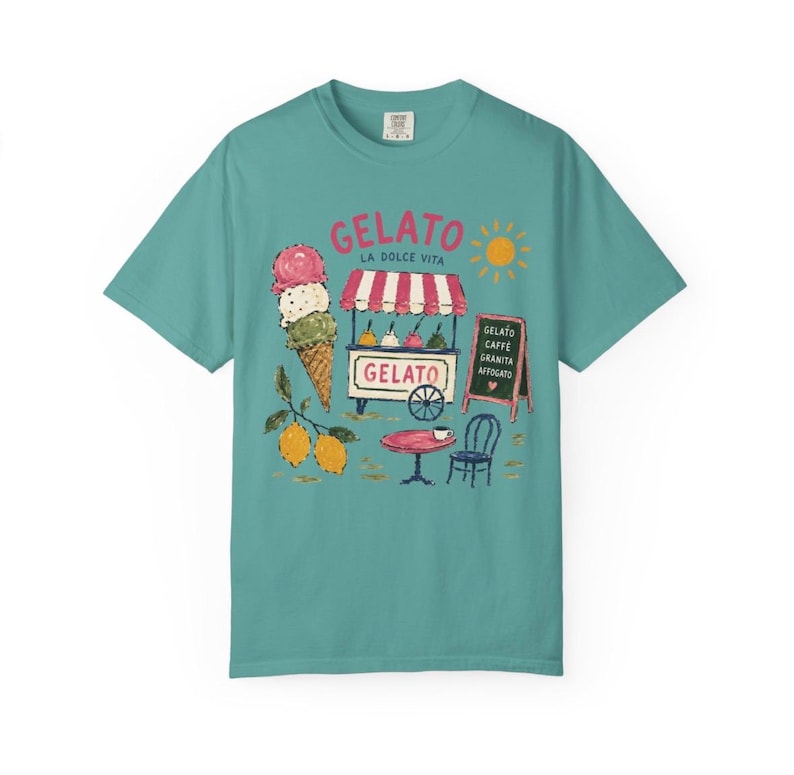Puede incluir: Camiseta color turquesa con un dise&ntilde;o de helado. El gr&aacute;fico incluye un cono de helado, un carrito de helado con la palabra "Gelato", una mesa, una silla y el texto "La Dolce Vita". Tambi&eacute;n se incluye un sol y limones.