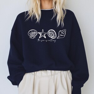 May include: A navy blue sweatshirt with a white graphic of seashells and a starfish. The text "The sea is calling" is printed below the graphic.