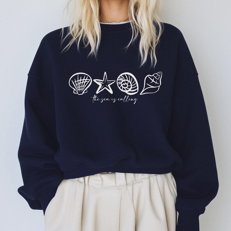 May include: A navy blue sweatshirt with a white graphic of seashells and a starfish. The text "The sea is calling" is printed below the graphic.