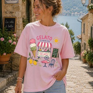 Puede incluir: Camiseta rosa con dise&ntilde;o de helado. El gr&aacute;fico incluye un carrito de helados, un cono de helado, limones y el texto "GELATO LA DOLCE VITA". Tambi&eacute;n hay una ilustraci&oacute;n de un sol. La camiseta la lleva una persona.