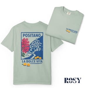 May include: Two light green t-shirts. One displays a graphic of Positano, Italy, with red flowers, blue buildings, and yellow lemons, with the text "LA DOLCE VITA." The other shirt shows the text "LA DOLCE VITA" and lemons. The brand name "ROSY" is at the bottom.