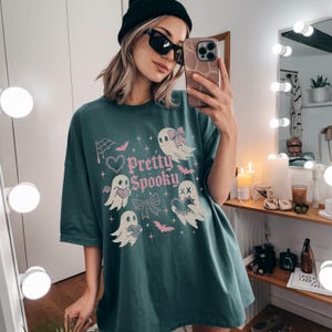 May include: Teal oversized t-shirt with a "Pretty Spooky" graphic. The design features cartoon ghosts, bats, bows, and a spiderweb in pink. The shirt has short sleeves and a relaxed fit.