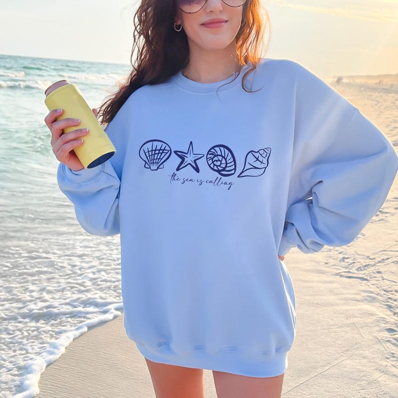 May include: Light blue sweatshirt with a graphic of seashells and a starfish. The text "The sea is calling" is printed below the graphic.