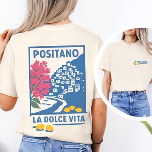 May include: Cream-colored t-shirt featuring a blue and white graphic of Positano, Italy, with pink flowers and yellow lemons. The text "POSITANO" and "LA DOLCE VITA" are displayed. A smaller version of the design is on the front.
