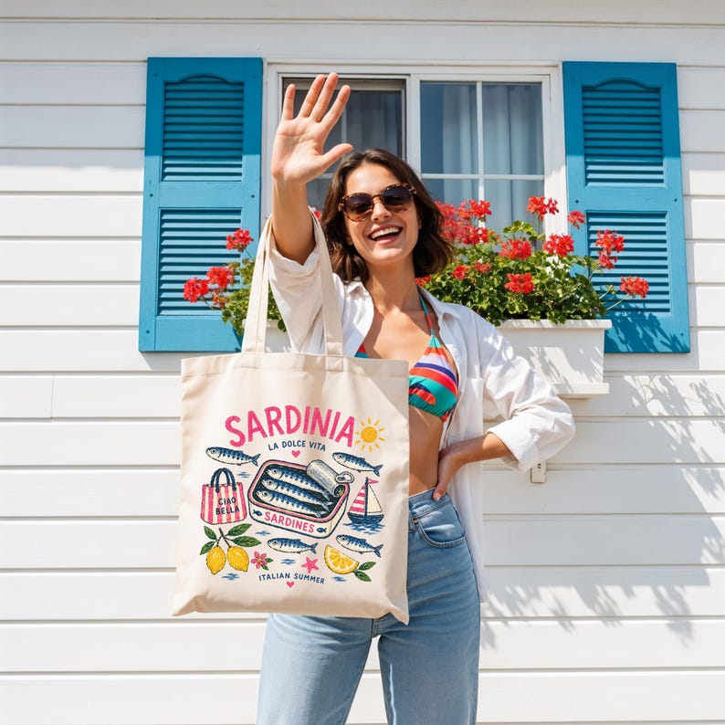 May include: A beige tote bag with the word "SARDINIA" in pink, featuring illustrations of sardines, lemons, and a sailboat. The bag also has the text "CIAO BELLA" and "ITALIAN SUMMER". The background includes a white building with blue shutters and red flowers.